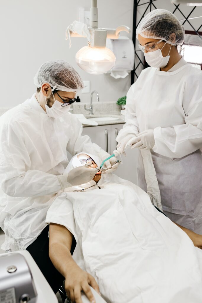 Dentysta w pełnym stroju ochronnym przeprowadza zabieg usuwania zęba u pacjenta leżącego na fotelu dentystycznym, z asystentką w tle.